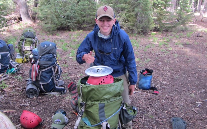 a person stands in front of a backpack resting on the ground and gives the camera a thumbs up